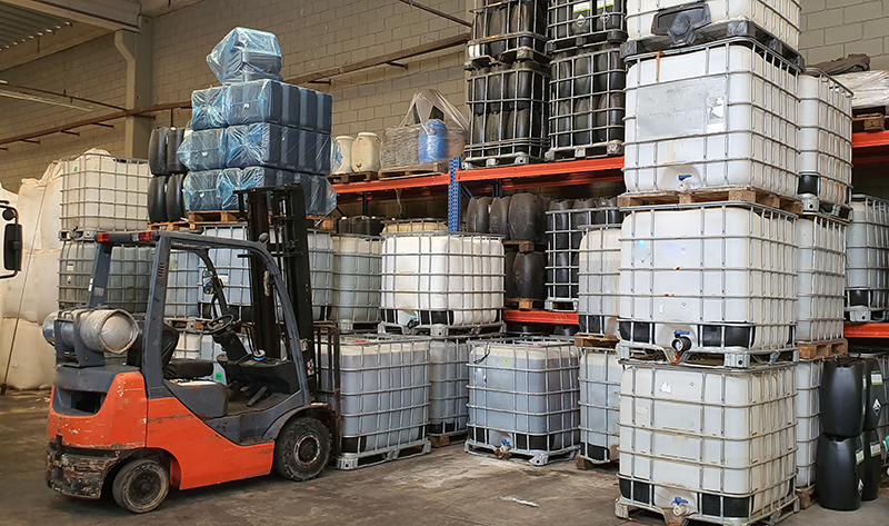 Een vorkheftruck tilt gestapelde Intermediate Bulk Containers (IBC's) in een industrieel magazijn.