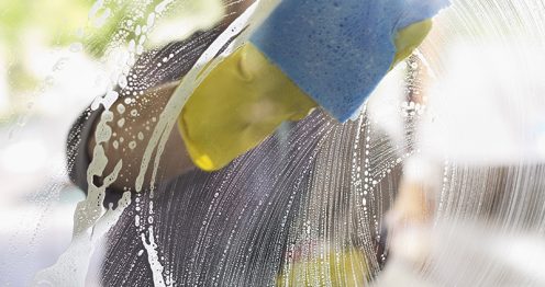 Een persoon wast een glazen venster met zeepachtig water en een blauwe spons, waarbij gele rubberen handschoenen worden gedragen.