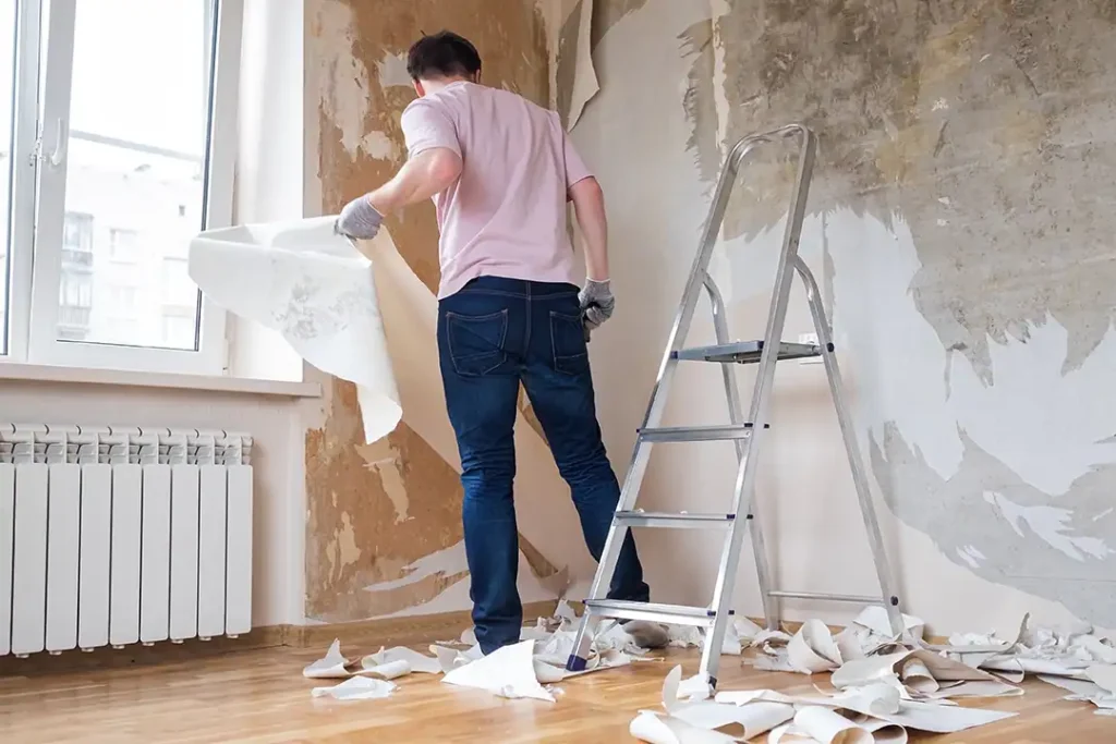 Ein Mann entfernt während einer Renovierung alte Tapeten von Hand von der Wand; daneben steht eine Leiter.