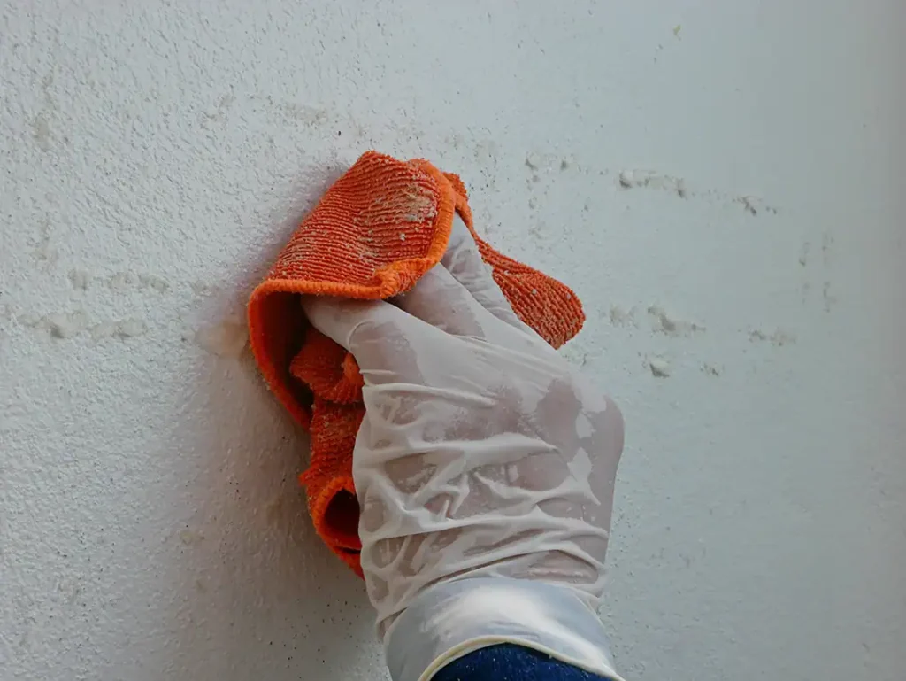 Eine Hand mit Kunststoffhandschuh entfernt mit einem orangefarbenen Tuch Klebstoffreste von einer weißen Wandoberfläche.