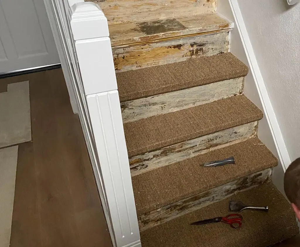 Eine Holztreppe, von der Teppich entfernt wird, mit sichtbaren Kleberesten sowie Werkzeugen wie Cuttermesser und Schaber auf den Stufen.
