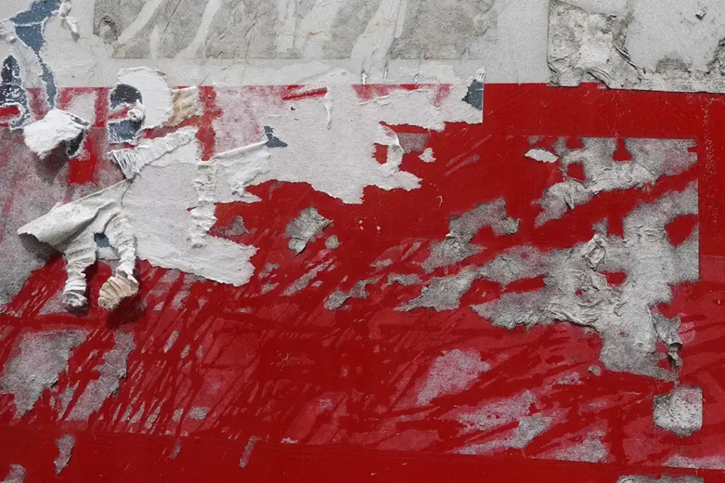 Eine Nahaufnahme hartnäckiger Klebereste auf einer roten Oberfläche mit teilweise abgerissenen und verblassten Papier- und Klebstoffschichten.