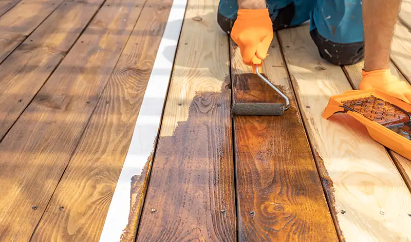 Person, die eine Holzterrasse mit Leinöl und einer Farbrolle behandelt.