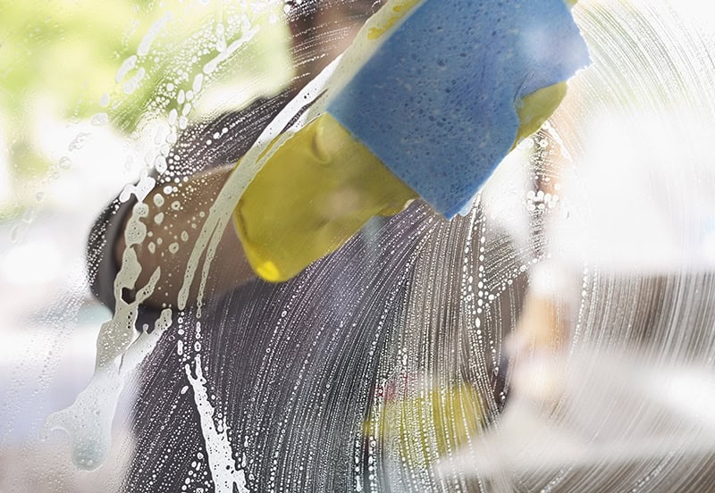 Eine Person mit gelben Gummihandschuhen reinigt ein Glasfenster mit Seifenwasser und einem blauen Schwamm.